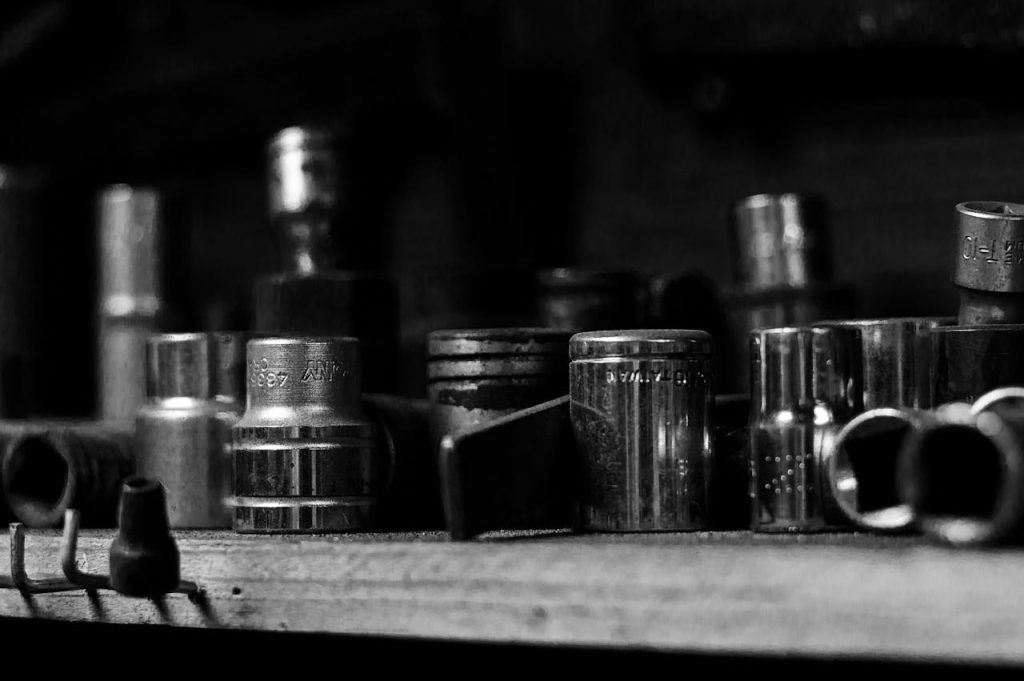 Black and white close-up of assorted metal socket heads on a workshop shelf.